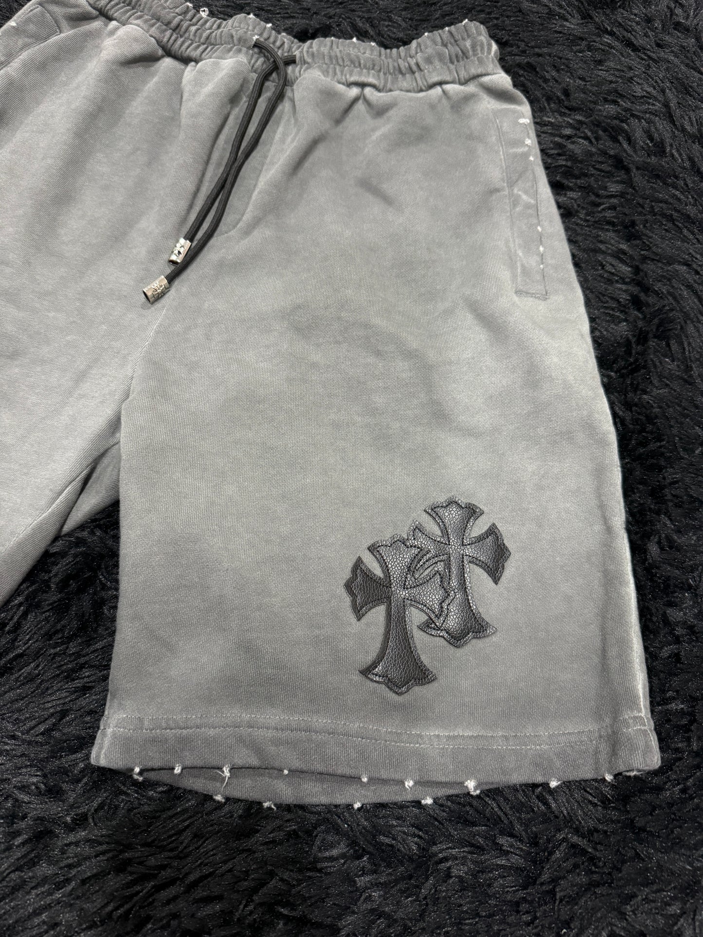 Gray Chrome Hearts shorts with a black cross design on a dark textured surface